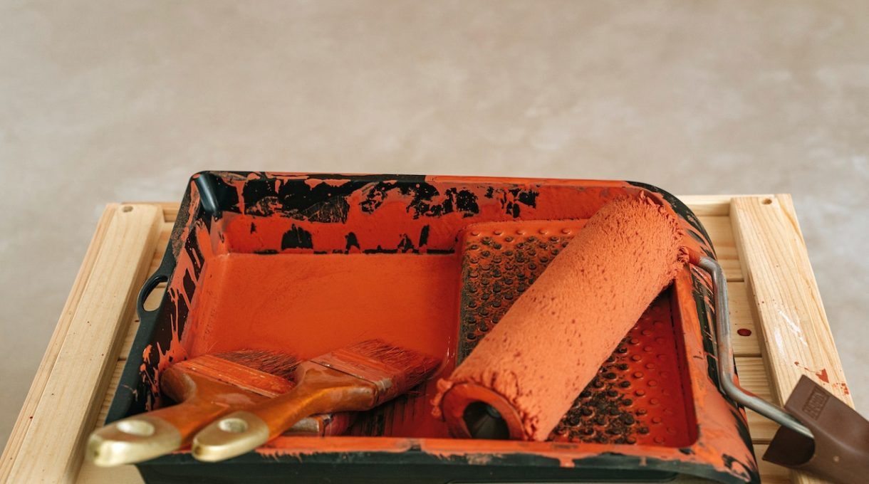Paint roller and brushes with orange paint on a plastic tray.